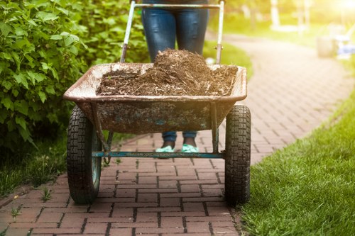 A person, whose upper body is not visible, is pushing a rusty metal wheelbarrow filled with dark soil or compost along a brick-paved pathway. The wheelbarrow has black rubber tyres and handles covered with foam grips. The pathway runs through a well-maintained garden area with lush green grass on one side and dense foliage or bushes on the other. In the background, a section of a paved outdoor space is visible, with sunlight filtering through the trees, creating a warm and inviting atmosphere. This scene reflects an outdoor activity involving garden waste or soil collection, consistent with rubbish removal services provided by Dalston House Clearance in London, suggesting a focus on garden clearance or waste disposal within an urban or suburban setting.