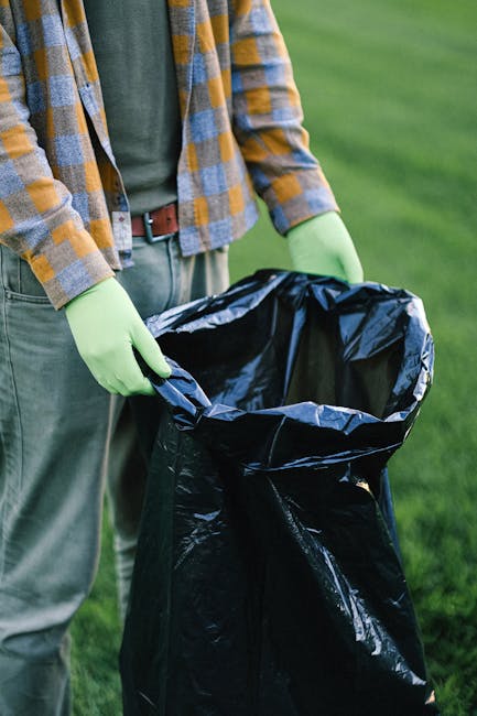 A row of large, rectangular wheelie bins positioned along a pavement beside a brick wall. The bins are made of durable plastic with a smooth finish, and each has a hinged lid on top. The bins are in t